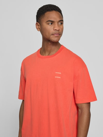 Męski model nosi T-Shirt z label-printem model 'JOEL' od Samsøe Samsøe, czysta bawełna, krój relaxed fit, okrągły dekolt, kolor pomarańczowy, długość tyłu w rozmiarze M: 71 cm, zbliżenie na górę ciała.
