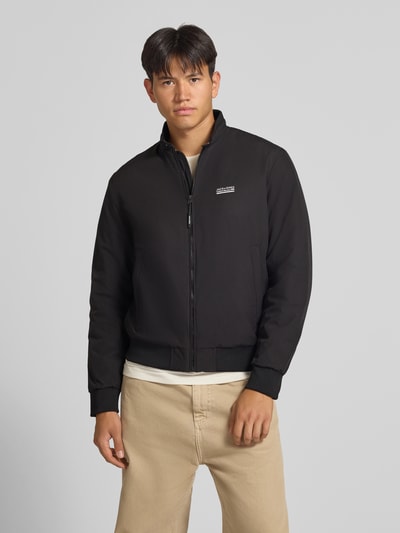 Mannenmodel draagt zwarte bomberjack met Logo Modell 'BROOKLYN' van JACK & JONES, regular fit, opstaande kraag, ritssluiting, ruglengte maat M 67 cm, schouderbreedte 49 cm, borstwijdte 59 cm, mouwlengte 66 cm, voorkant volledige lichaamsopname.