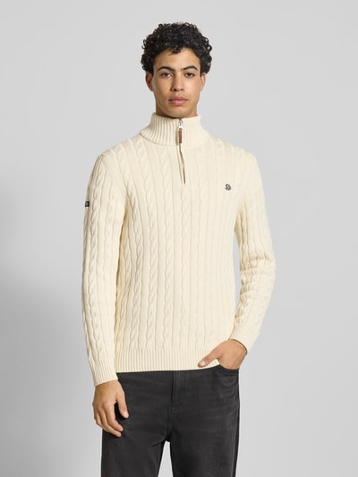 Männermodel trägt Strickpullover aus reiner Baumwolle von Superdry in Creme mit Zopfmuster, Troyer-Kragen, Label-Detail, gerippte Abschlüsse, enganliegende Passform, Rückenlänge 70 cm, Ärmellänge 71 cm; Ganzkörperansicht von vorne.