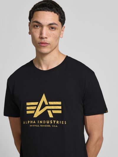 Männermodel trägt Herren T-Shirt mit Label-Detail Modell 'Basic' von Alpha Industries in Schwarz mit Goldprint, Reine Baumwolle, Gerader-Schnitt, Rundhalsausschnitt, Rückenlänge bei Größe M: 67 cm, Nahaufnahme Oberkörper.