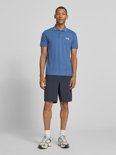 Męski model nosi Regular Fit Poloshirt mit Logo-Print od EA7 Emporio Armani w niebieskim kolorze, bawełna z domieszką elastanu, krój regular fit, nadruk logo, kołnierzyk polo, krótkie rękawy, elastyczne mankiety, długość tyłu w rozmiarze M: 69 cm; pełny widok z przodu.