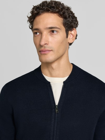 Männermodel trägt Slim Fit Strickjacke aus Baumwoll-Mix mit Stehkragen von Christian Berg Men, dunkle Farbe, Nahaufnahme Oberkörper. Kontext: Herren Strickjacke, Zweiwege-Reißverschluss, langärmliges Design, gerippte Abschlüsse.