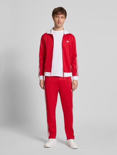 Männermodel trägt Regular Fit Sweatjacke mit Stehkragen Modell 'Classic' von adidas Originals aus Baumwoll-Mix in Rot mit Logo-Stitching, Zweiwege-Reißverschluss und Eingrifftaschen, elastische Abschlüsse, Ganzkörperansicht von vorne.
