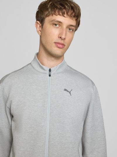Mężczyzna nosi bluza rozpinana 'Sweatjacke mit Stehkragen und Logo' od PUMA PERFORMANCE, mieszanka materiałów z dodatkiem lyocellu, krój regular fit, stójka, logo, zamek błyskawiczny, długość tyłu w rozmiarze M: 71 cm, długość rękawów w rozmiarze M: 70 cm, jasnoszara, tekstura gładka, prosty fason, widoczny detal z logo, tło jednokolorowe, zbliżenie na górną część ciała, widok z przodu.