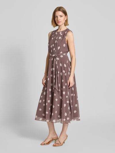 Vrouwenmodel draagt Midikleid met gelegde plooien van Christian Berg Cocktail in bruin met witte stippen, getailleerd, ronde hals, afneembare stoffen band, mouwloos design, onderrok, ruglengte bij maat 36: 120 cm, borstwijdte bij maat 36: 45 cm. Ganzkörperansicht van voren.