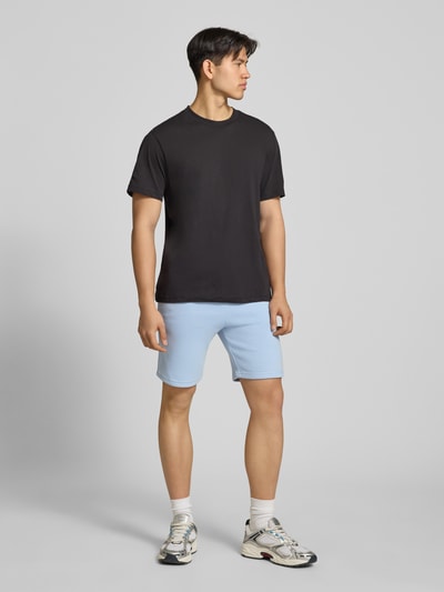 Mannelijk model draagt Shorts mit Eingrifftaschen und Logo-Print van Alpha Industries in lichtblauw katoenmix, relaxed fit, elastische band met trekkoord, steekzakken, logoprint, achterzak, bandwijdte maat M 39 cm, binnenbeenlengte 24 cm, geheelvooraanzicht.