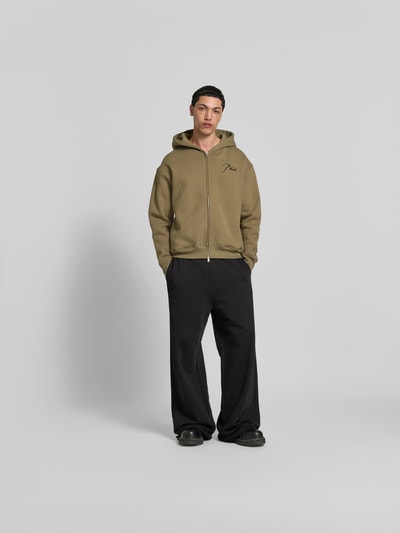Männermodel trägt Oversized Sweatjacke aus Baumwolle von RHUDE in Beige mit Zweiwege-Reißverschluss und Label-Stitching, elastische Abschlüsse, Rückenlänge 70 cm, Schulterbreite 62 cm, Ärmellänge 66 cm, Ganzkörperansicht von vorne.
