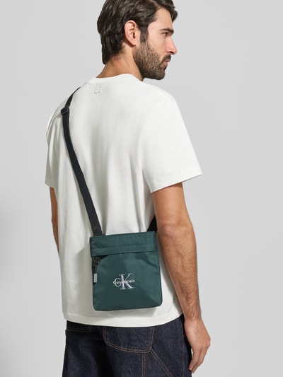 Männermodel trägt Crossbody Bag mit Label-Stitching von CK Calvin Klein in Dunkelgrün mit schwarzem Riemen, Hauptfach mit Reißverschluss, Ganzkörperansicht Rückansicht.