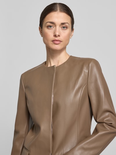 Kobiece model prezentuje kurtkę Slim Fit Jacke in Leder-Optik model Doma od STAND STUDIO, materiał skóropodobny, krój slim fit, okrągły dekolt, kieszenie z patką, długość tyłu w rozmiarze 36: 70 cm, szerokość ramion w rozmiarze 36: 40 cm, szerokość w rozmiarze 36: 46 cm, długość rękawów: 64 cm, jasnobrązowy, gładka tekstura; zbliżenie na górną część ciała z przodu.