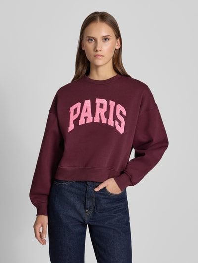Vrouwenmodel draagt Relaxed Fit Sweatshirt uit katoenmix Model 'Tatum' van JJXX, bordeaux met roze statementpatch, ronde hals, ribboordjes, met lange mouwen, verlaagde schouders, detailopname borst, dames sweatshirt, katoenmix, relaxed fit. Ganzkörperansicht von voren.
