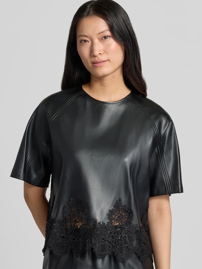 Frauenmodel trägt Blusenshirt in Leder-Optik mit Spitzenbesatz Modell 'LONDON' von SUNCOO PARIS in Schwarz, Regular Fit, Nahaufnahme Oberkörper.