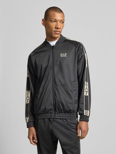 Männermodel trägt schwarze Trainingsjacke mit Zweiwege-Reißverschluss von EA7 Emporio Armani, Regular-Fit, Logo, Stehkragen, Eingrifftaschen, Ganzkörperansicht von vorne.