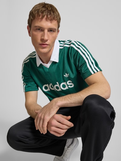 Männermodel trägt Trikot mit Umlegekragen und Label-Print von adidas Originals in Grün mit weißen Details, Regular Fit, Nahaufnahme Oberkörper. Rückenlänge bei Größe M: 73 cm.