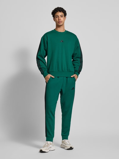 Männermodel trägt grüne Sweatpants mit elastischem Bund von adidas Sportswear, Baumwoll-Mix mit Viskose-Anteil, Tunnelzug, Lockerer-Schnitt, Label-Stitching, Ganzkörperansicht von vorne.