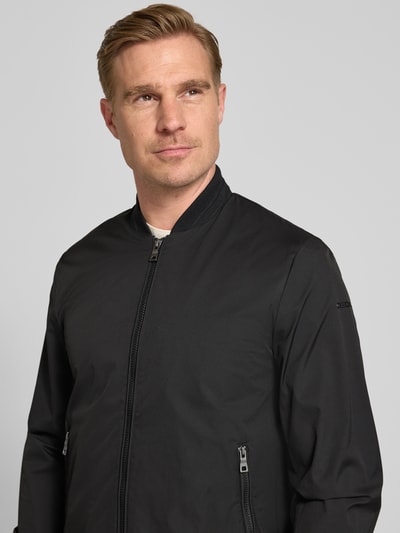 Mannenmodel draagt Bomberjacke mit Reißverschlusstaschen van Geox in zwart met opstaande kraag, regular fit, ritszakken, ritssluiting. Nahaufnahme bovenlichaam. Context: heren bomberjack van Geox, regular fit, opstaande kraag, ritssluiting, ritszakken, ruglengte bij maat M: 68 cm, schouderbreedte bij maat M: 46 cm, borstwijdte bij maat M: 45 cm, mouwlengte bij maat M: 64 cm.