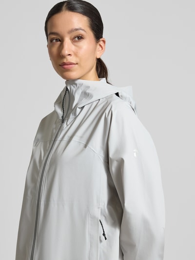 Frauenmodel trägt Mantel mit Zweiwege-Reißverschluss und Kapuze von PEAK PERFORMANCE in hellem Grau, glatte Textur, Nahaufnahme Oberkörper. Relaxed Fit, Modell TREELINE HIPE® 2.5L SHELL, Logo sichtbar, Kapuze vorhanden.