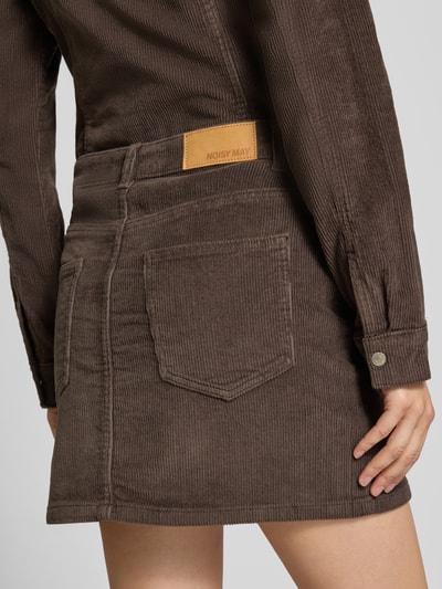 Frauenmodel trägt Minirock aus Baumwoll-Mix in Cord-Optik Modell 'Yolanda' von NOISY MAY®, braun, 5-Pocket-Design, Detailaufnahme Rückansicht. Körpernahe Passform, Label-Patch sichtbar, Struktur mit feiner Rippenoptik, Kontext: elastischer Baumwoll-Mix, Knopf- und Reißverschluss, Bundweite S 39 cm, Gesamtlänge S 41 cm.