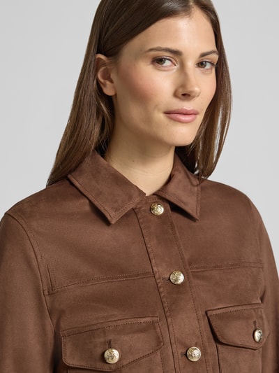 Kobiece model prezentuje kurtkę 'Jacke mit Umlegekragen' od Oui, elastyczna mieszanka materiałów, prosty krój, wykładany kołnierz, zapięcie na guzik, nakładane kieszenie, długość tyłu w rozmiarze 36: 49 cm, szerokość ramion w rozmiarze 36: 39 cm, szerokość klatki piersiowej w rozmiarze 36: 43 cm, długość rękawów w rozmiarze 36: 61 cm, kolor brązowy, tekstura gładka, detaliczne złote guziki. Zbliżenie górnej części.
