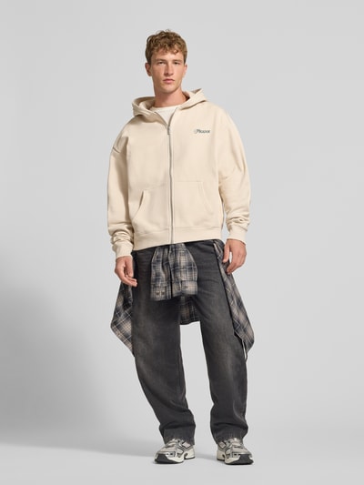 Männermodel trägt Relaxed Fit Sweatjacke aus Baumwoll-Mix Modell 'Carloforte' von PEGADOR in Beige mit Logo-Stickerei, geteilte Kängurutasche, langärmlig, gerippte Abschlüsse, überschnittene Schultern, Rückenlänge 69 cm, Ärmellänge 68 cm, kombiniert mit dunkler Hose, Ganzkörperansicht von vorne.