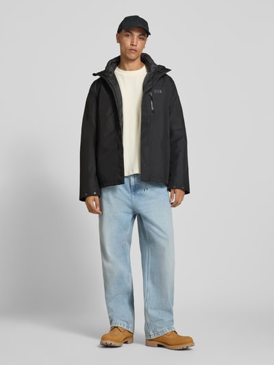 Männermodel trägt Regular Fit Jacke mit Logo-Print Modell 'Juell' von Helly Hansen in Schwarz, Kapuze, Taschen, Ganzkörperansicht von vorne.