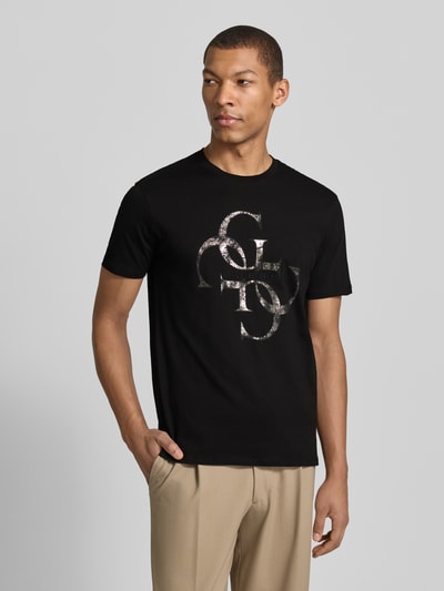 Męski model nosi T-Shirt mit Label-Detail od Guess, czarna bawełna, prosty krój, nadruk z logo, długość tyłu w rozmiarze M: 69 cm, całkowity widok z przodu