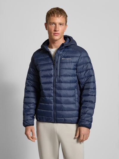 Mannenmodel draagt Steppjacke met Kapuze und Logo van CHAMPION in blauw met rits, steekzakken, logo, regular fit, vooraanzicht bovenlichaam.