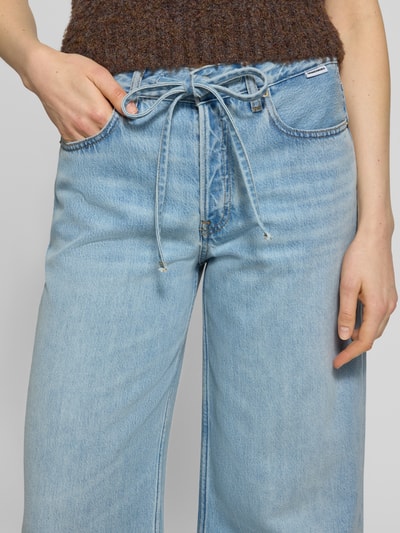 Kobiece modelka nosi Loose Fit Jeans aus Baumwoll-Lyocell-Mix Modell 'ASTRAEAAS' von Armedangels, stylizowana na denim, krój loose fit, średni stan, naszywka z logo, szlufki na pasek, zapięcie na guzik i ukryty zamek błyskawiczny, 5 kieszeni, materiał: mieszanka bawełny i lyocellu, jasnoniebieski, miękka tekstura, zbliżenie na dół tułowia