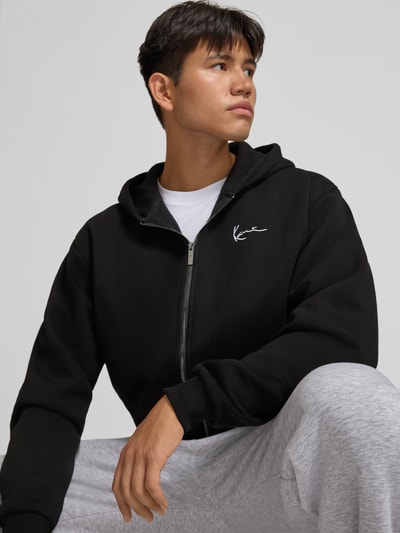 Männermodel trägt schwarze Sweatjacke mit Label-Stitching und Känguru-Tasche von Karl Kani, Baumwoll-Mix, Nahaufnahme Oberkörper.