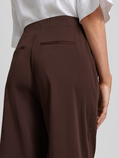 Kobiece model nosi spodnie materiałowe z mieszanki bawełny z dodatkiem streczu, krój loose fit, model 'Mucia Base' od OPUS PANTS, elastyczny pas, wpuszczane kieszenie z przodu, kieszenie z wypustką z tyłu, kolor ciemnobrązowy, długość wewnętrzna nogawki 80 cm, szerokość w rozmiarze 36: 31 cm, detaliczne ujęcie tylnej części.
