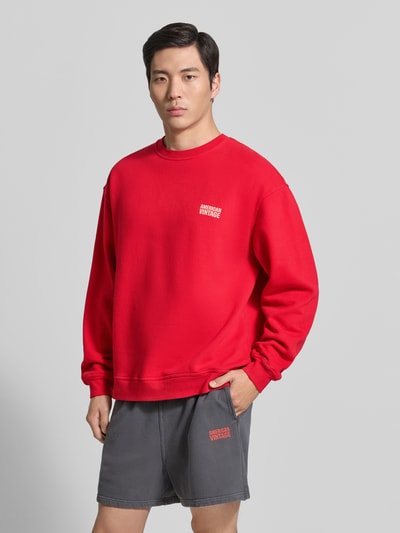 Męskimodel nosi czerwoną bluzę z logo, krój relaxed fit, czysta bawełna, okrągły dekolt, model 'PLIZZY' od American Vintage, długość tyłu 66 cm, długość rękawów 60 cm, zdjęcie całej sylwetki z przodu.