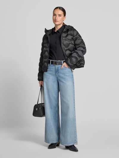 Frauenmodel trägt Steppjacke mit Kapuze und Reißverschlusstaschen von Marc Cain, czarny kolor, krój regular fit, kaptur z tunelem, zamek błyskawiczny dwustronny, kieszenie zapinane na zamek błyskawiczny, długość tyłu w rozmiarze 36: 58 cm, szerokość ramion w rozmiarze 36: 43 cm, szerokość w rozmiarze 36: 38 cm, długość rękawów w rozmiarze 36: 62 cm, całkowity widok z przodu.