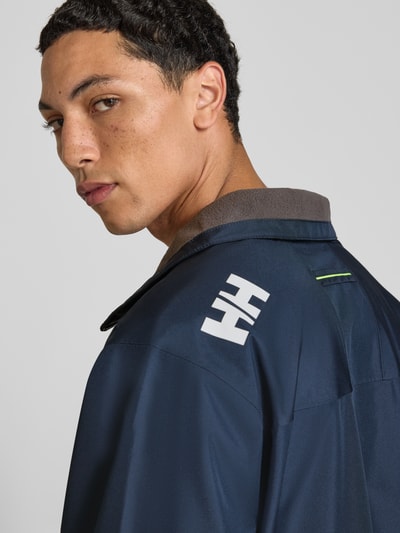Männermodel trägt Regular Fit Jacke mit Stehkragen Modell 'Crew' von Helly Hansen in Navy mit Logo-Print, Nahaufnahme Oberkörper.