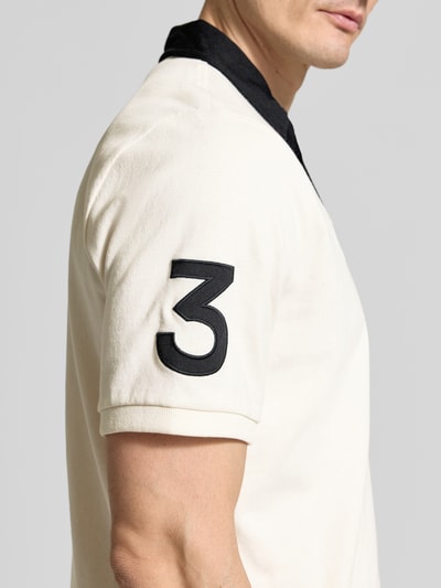 Mężczyzna nosi Regular Fit Poloshirt aus reiner Baumwolle von Polo Ralph Lauren, kremowy kolor, czarne detale, krój regular fit, logo na rękawie, materiał bawełniany, zbliżenie na górną część ciała