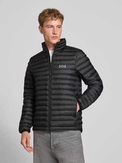 Männermodel trägt schwarze Steppjacke mit Label-Print von Helly Hansen, durchgehender Reißverschluss, elastische Ärmel, Ganzkörperansicht von vorne