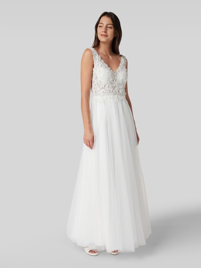 Modelka nosi Brautkleid mit Ziersteinen und floraler Spitze od Mascara, taliowany krój, dekolt w serek, szerokie ramiączka, koronka kwiatowa, ozdobne kamienie, półprzezroczysty materiał wierzchni, kryjąca halka, zapięcie na haftkę i wszyty zamek błyskawiczny, długość tyłu w rozmiarze 36: 150 cm, szerokość klatki piersiowej w rozmiarze 36: 39 cm, zdjęcie całej sylwetki z przodu.