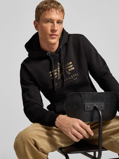Männermodel trägt Hoodie mit Logo und Känguru-Tasche von Alpha Industries, schwarz-gold, Baumwoll-Mix, Regular-Fit, Nahaufnahme Oberkörper.