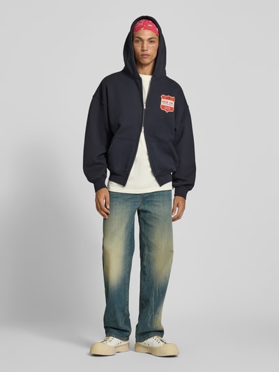 Mannelijk model draagt Oversized Sweatjacke aus Baumwolle van RHUDE in donkerblauw met labelprint, kangoeroezak, puur katoen, tweewegritssluiting, ruglengte maat M 64 cm, schouderbreedte 45 cm, mouwlengte 56 cm, Ganzkörperansicht van voren.