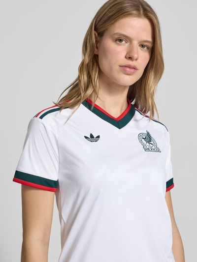 Kobiece model nosi Mexiko Auswärtstrikot 2026 od adidas Sportswear, biały z wieloma kolorami, krój regular fit, faktura, aplikacja z logo, wyhaftowane logo, paski w kontrastowym kolorze, dekolt w serek, krótki rękaw, zakończenie rękawów prążkowane i z lamówką, szybkoschnący, materiał odprowadzający wilgoć, zbliżenie na górną część ciała.