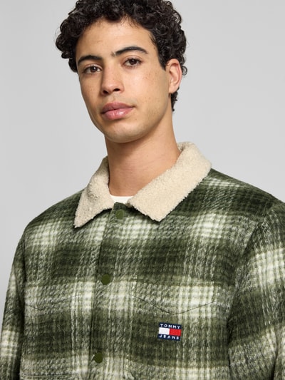 Mężczyzna nosi Oversized Hemdjacke mit Allover-Muster von Tommy Jeans, efekt: wzór na całej powierzchni, wykładany kołnierz, listwa z zatrzaskami, naszywka z logo, długi rękaw z zakończeniem na zatrzask, wpuszczane kieszenie boczne i kieszeń na piersi, kolor zielony z beżowym kołnierzem, struktura materiału miękka, Nahaufnahme górnej części ciała.