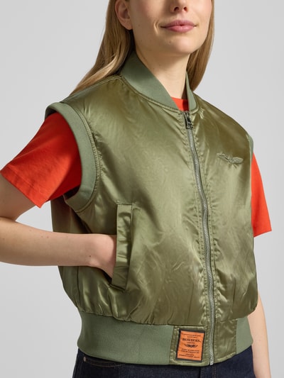 Frauenmodel trägt Weste mit Reißverschluss und Label-Detail von Original Bombers in Oliv, Relaxed-Fit, Eingrifftaschen, Nahaufnahme Oberkörper. Rückenlänge 52 cm, Schulterbreite 44 cm bei Größe S.