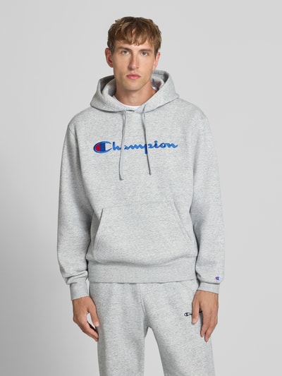 Männermodel trägt Hoodie mit Label-Stitching von Champion in Hellgrau, Baumwoll-Mix, lockerer Schnitt, Ganzkörperansicht von vorne.