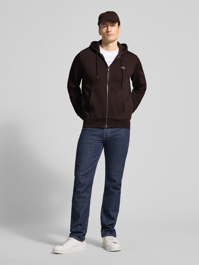 Męski model nosi Regular Fit Sweatjacke aus Baumwoll-Mix von Lacoste, jednokolorowy, krój regular fit, zamek błyskawiczny, naszywka z logo, kaptur z tunelem, długi rękaw, zakończenia prążkowane, przednie kieszenie, dzianina dresowa, mieszanka bawełny. Ganzkörperansicht von vorne.