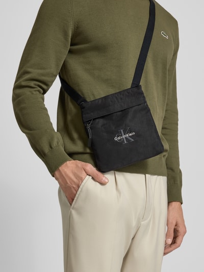 Männermodel trägt Crossbody Bag mit Label-Stitching von Calvin Klein in Schwarz, rechteckige Form, strukturierte Oberfläche, Nahaufnahme Oberkörper. Hauptfach mit Reißverschluss, verstellbarer Schulterriemen, etwa 21 x 21 x 3 cm, Riemen 80 cm lang.