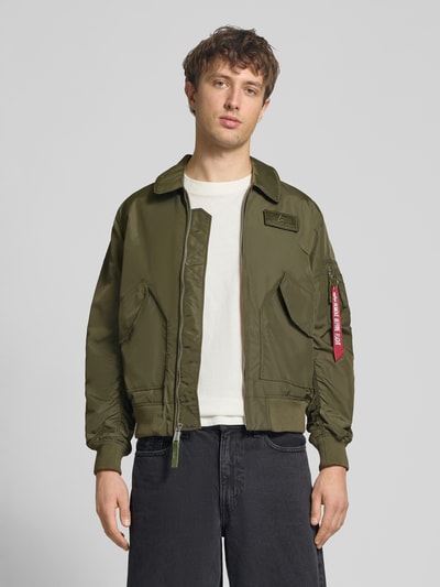 Männermodel trägt Regular Fit Bomberjacke mit Logo-Patch von Alpha Industries in Olivgrün, mit Umlegekragen und aufgesetzten Taschen, Ganzkörperansicht von vorne.