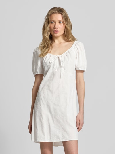 Frauenmodel trägt A-Linien Umstandskleid aus reiner Baumwolle Modell 'JUTTY' von Vero Moda in Optik: Unifarben mit Struktur-Muster, Mini-Länge, Karree-Ausschnitt mit Binde-Detail, Ballonärmel und elastischem Ärmelabschluss, Ganzkörperansicht von vorne.
