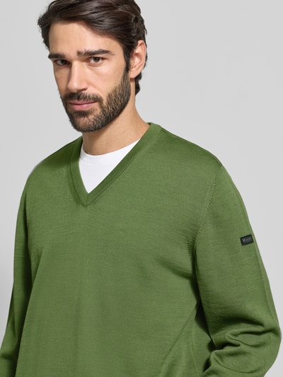 Männermodel draagt Strickpullover aus Schurwolle mit V-Ausschnitt van MAERZ Muenchen in groen, regular fit, labeldetail, Nahaufnahme Oberkörper.