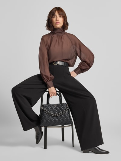 Frauenmodel trägt schwarze Hose, braune Bluse, schwarze Stiefeletten, Handtasche in Leder-Optik Modell 'Maxine' von GUESS auf Stuhl, Ganzkörperansicht von vorne. Damen Handtasche von GUESS mit geteiltem Hauptfach, Reißverschluss, abnehmbarem Schulterriemen, Logo-Detail, Maße 36x24x12 cm, Schulterriemen 120 cm.