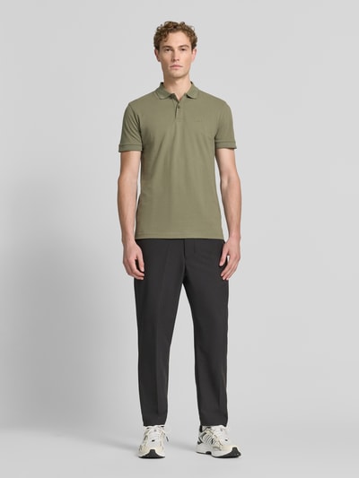 Męski model nosi Regular Fit Poloshirt aus reiner Baumwolle Modell 'PIO 1' von BOSS Green, oliwkowa koszulka polo, czarne spodnie, jasne sneakersy, stonowane kolory, gładka tekstura, Ganzkörperansicht von vorne.