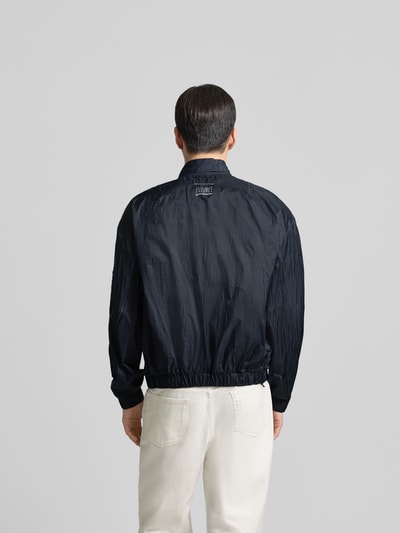 Männermodel trägt Relaxed Fit Jacke mit Logo-Print von MM6 Maison Margiela in Dunkelblau, elastische Abschlüsse, Rückenansicht, Ganzkörperansicht Rückansicht.