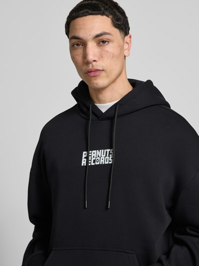 Męski model nosi Oversized Hoodie aus Baumwoll-Mix Modell 'PEANUTS BACK' von Only & Sons, czarny, nadruk peanuts®, dzianina dresowa, kieszeń kangurka, kaptur z tunelem, długi rękaw, zakończenia prążkowane, krój oversized, mieszanka bawełny, kolor jednolity, zbliżenie na górną część ciała, ujęcie z przodu